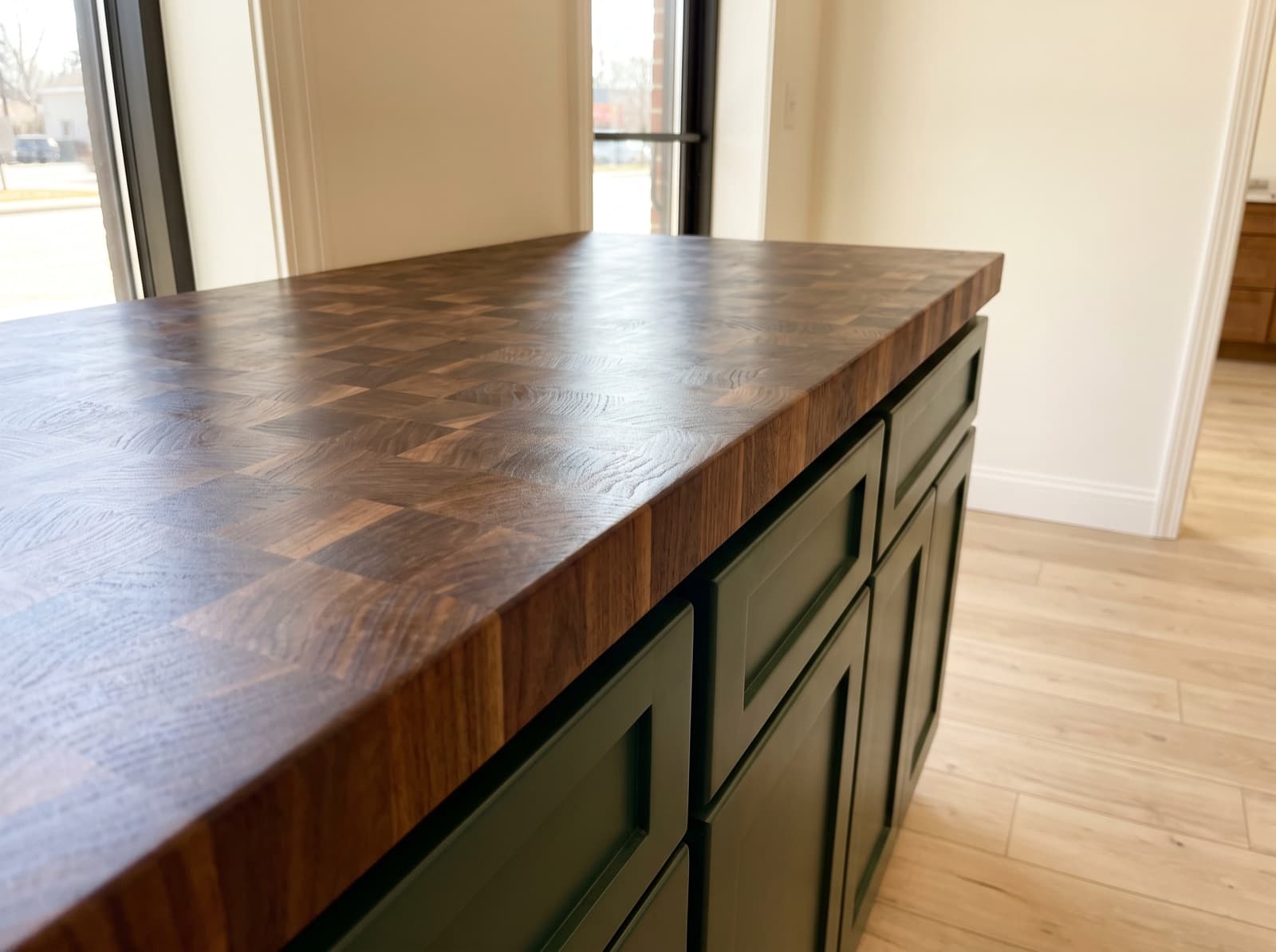 A freshly oiled end-grain walnut island top seen close along its surface, the chequerboard of block ends running to a thick square edge above deep green shaker cabinets.