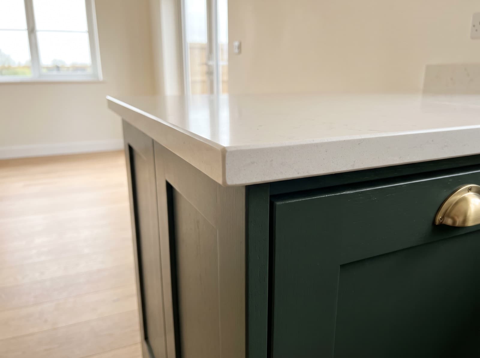 The corner of a newly fitted pale speckled quartz countertop with a plain eased edge, overhanging deep green shaker cabinets with a brass cup handle, in an empty kitchen lit from a side window.
