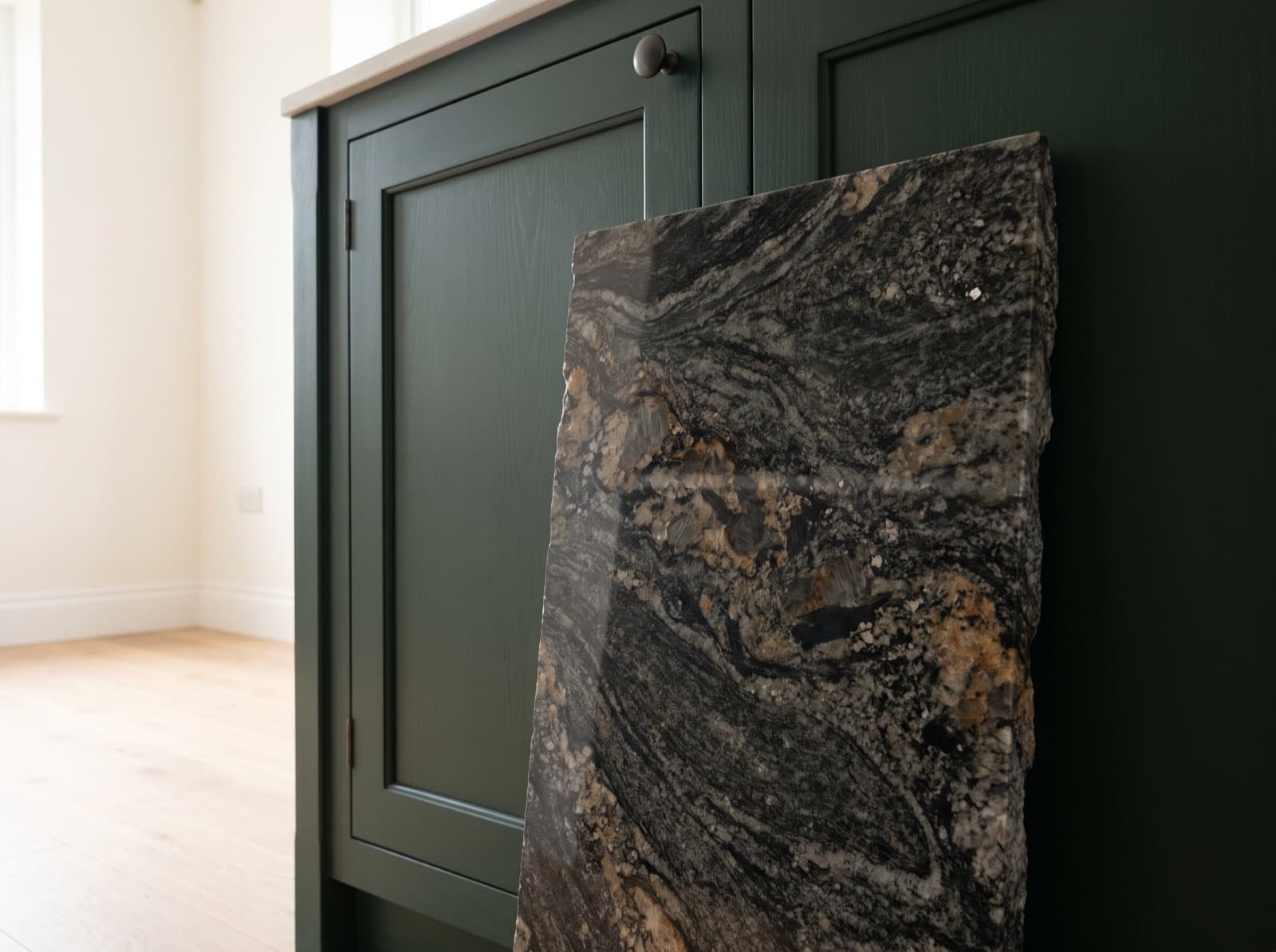 A granite offcut with a rough unfinished quarry edge and heavy black, gold and grey movement, leaning against a deep green cabinet door in an otherwise empty room.