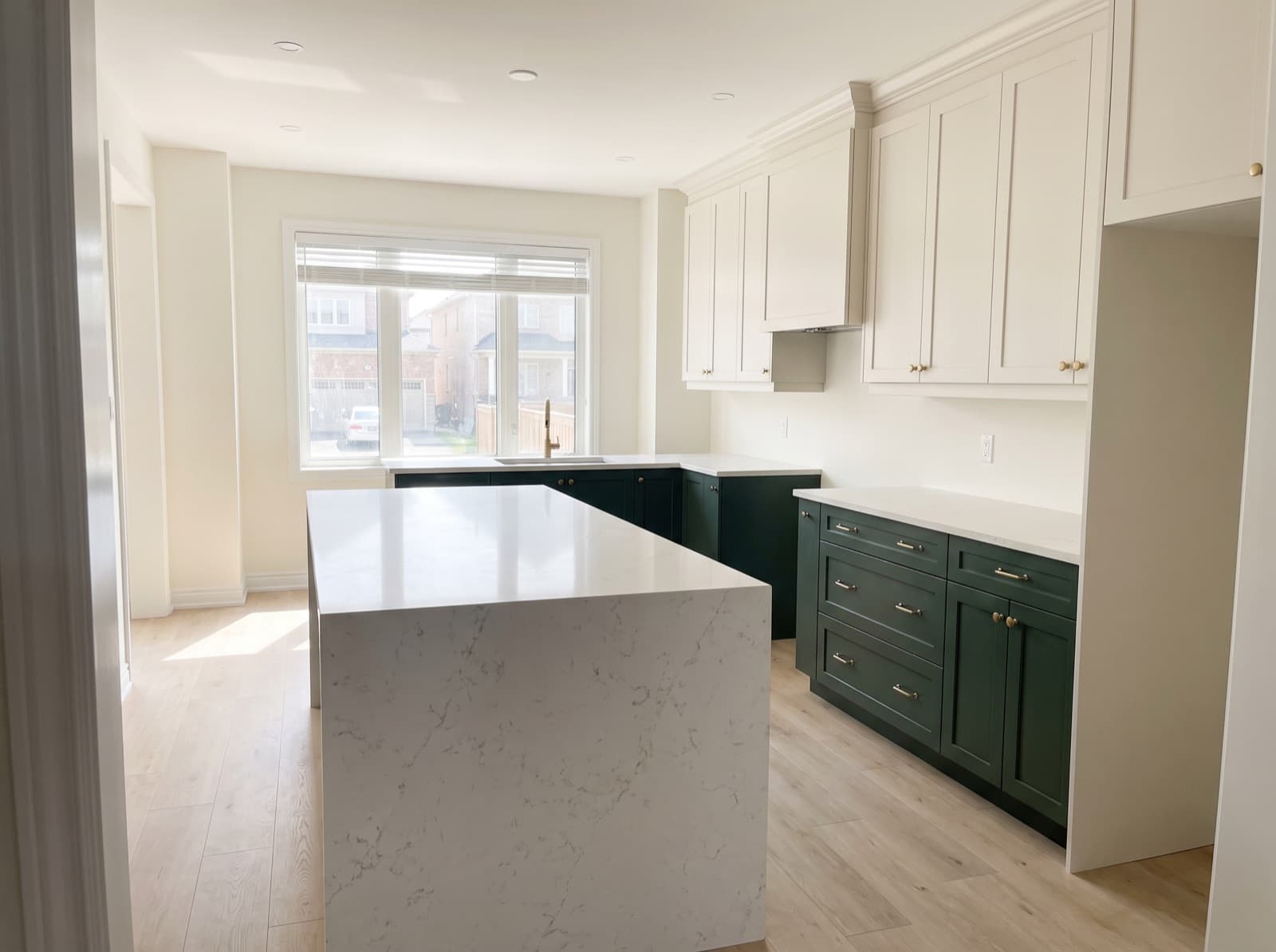 A finished but unfurnished kitchen seen from the doorway: deep green base cabinets, cream wall cabinets, and a white veined quartz island whose stone is carried down the end to the floor.