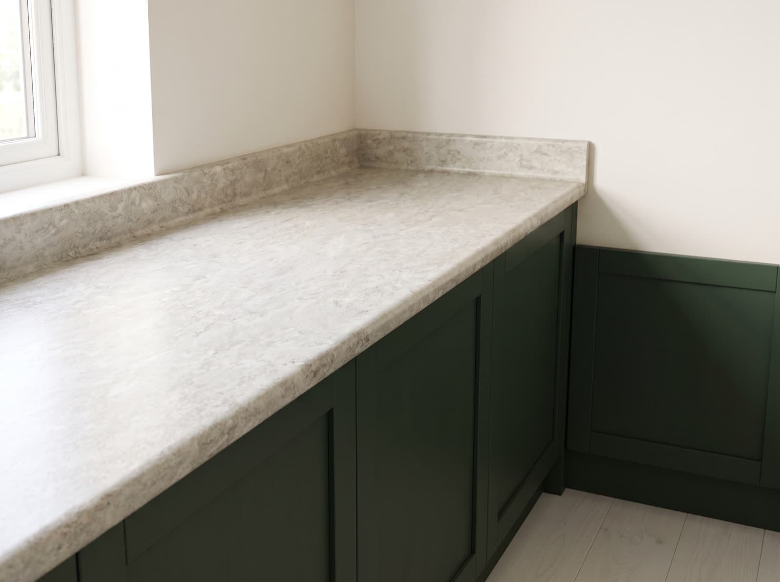 A stone-look laminate counter running into a corner under a window, with a rolled post-formed front edge and a coved upstand carried up from the same piece, on deep green cabinets.