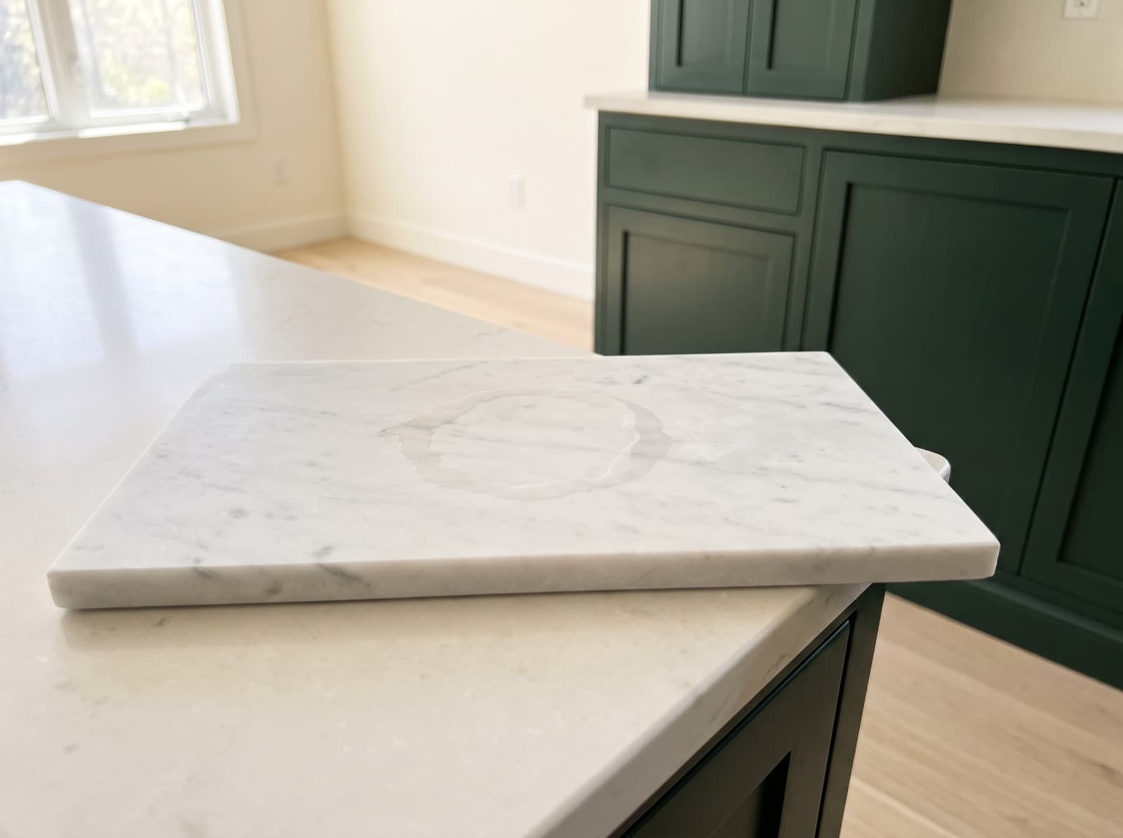 A rectangular white Carrara marble sample lying on a pale counter with a ring of water sitting on its surface, deep green cabinets and a pale counter run behind it.