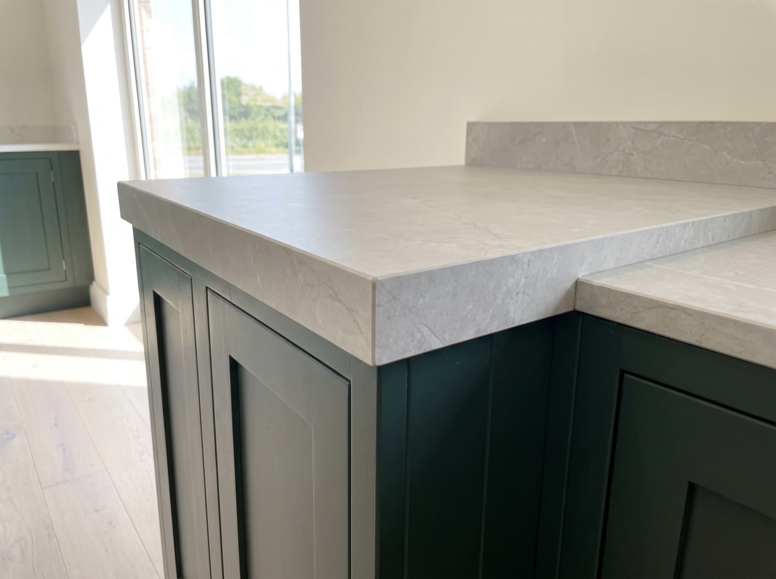The corner of a grey veined porcelain countertop, its front edge mitred and built up to full countertop thickness, on deep green cabinets with a matching porcelain upstand behind.