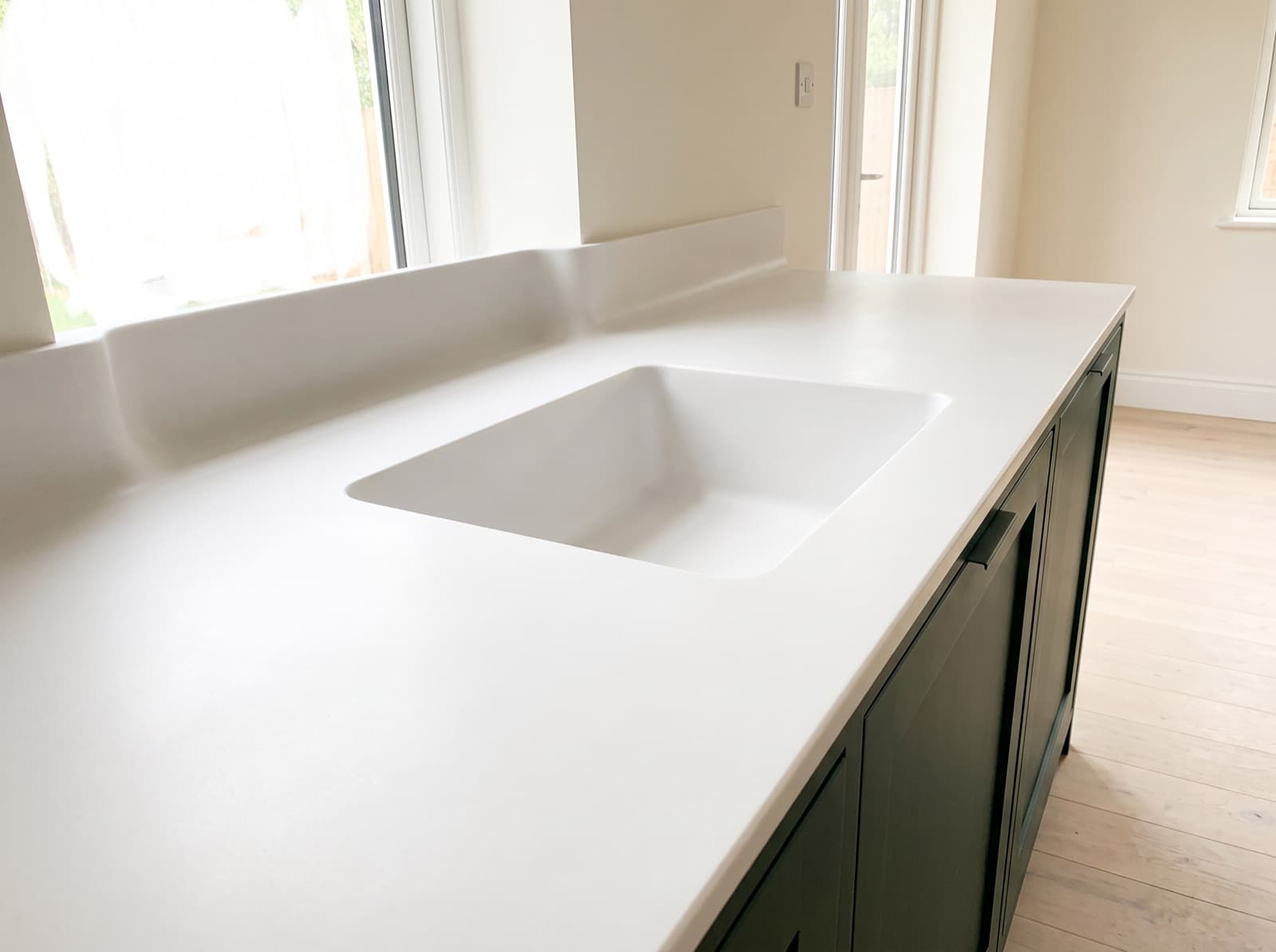 A matte white solid surface countertop whose rectangular sink bowl and coved upstand are formed from the same sheet, with no rim, seal or visible joint anywhere, above dark green cabinets.