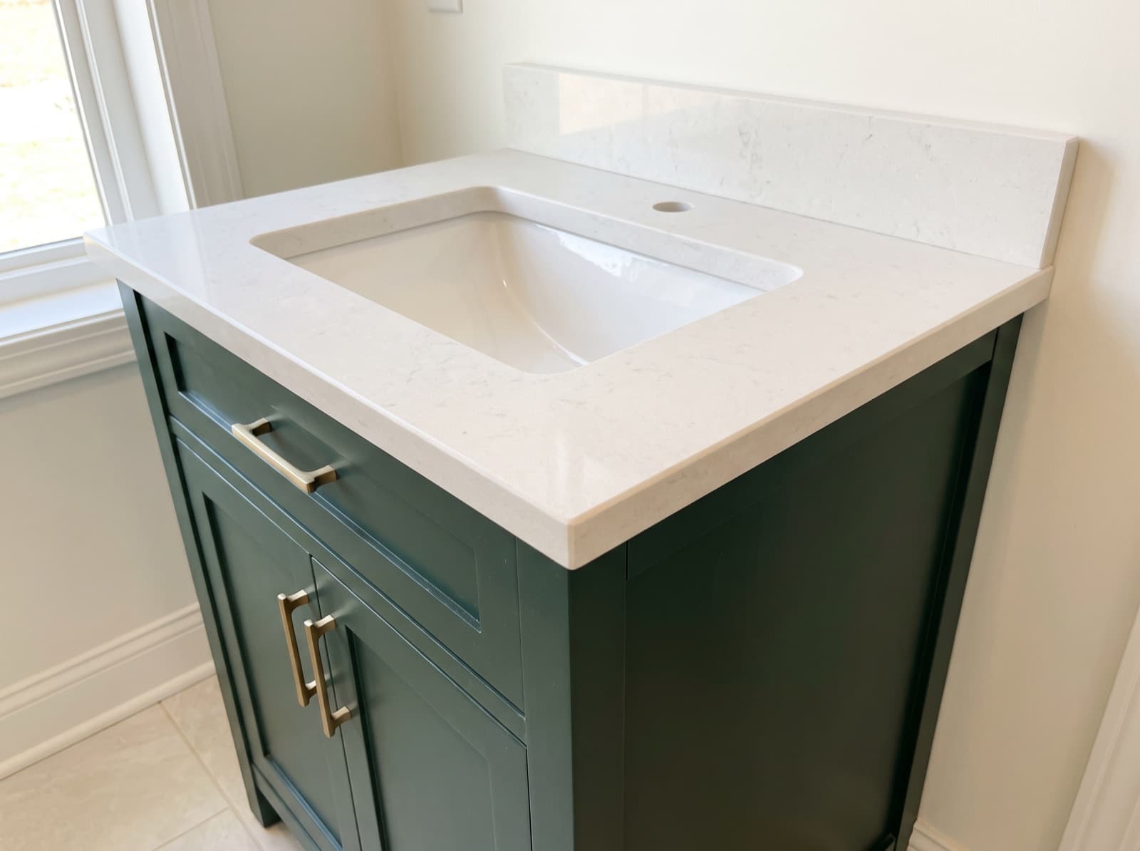A pale quartz vanity top with a rectangular undermount sink, a single tap hole and a matching upstand, sitting on a deep green vanity cabinet with brushed brass bar handles.